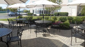 Un área de patio al aire libre con mesas y sillas redondas, protegida por grandes sombrillas, frente a un edificio de diseño moderno.