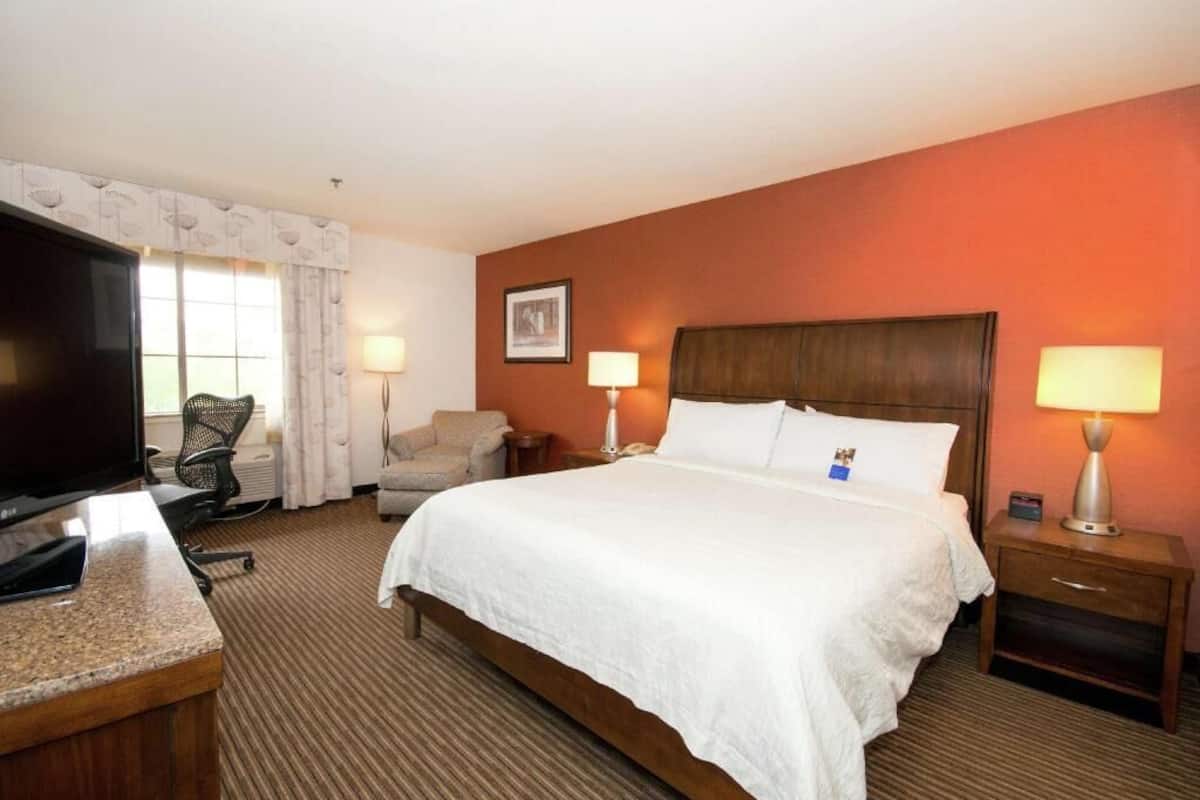 A hotel room with a bed, a desk with a flat-screen TV, two bedside lamps, a chair, and a framed picture on the wall.