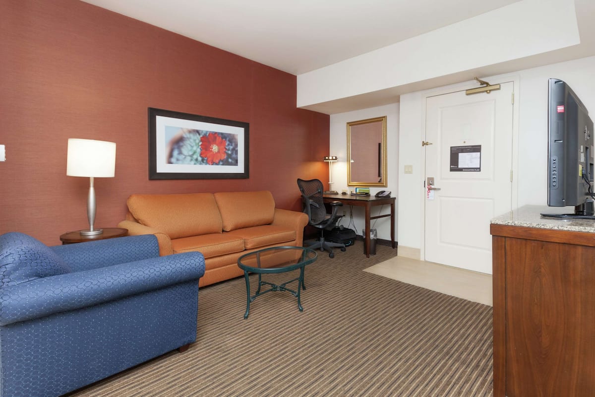A hotel room with a blue sofa, an orange armchair, a small round table, a desk with a chair, a television, and a door with a sign.