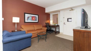 A hotel room with a blue sofa, an orange armchair, a small round table, a desk with a chair, a television, and a door with a sign.