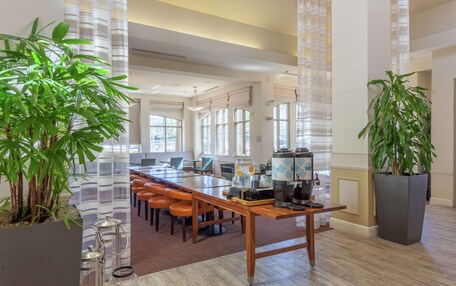 A spacious hotel lobby with a long wooden reception desk, multiple seating areas, large windows, and potted plants.