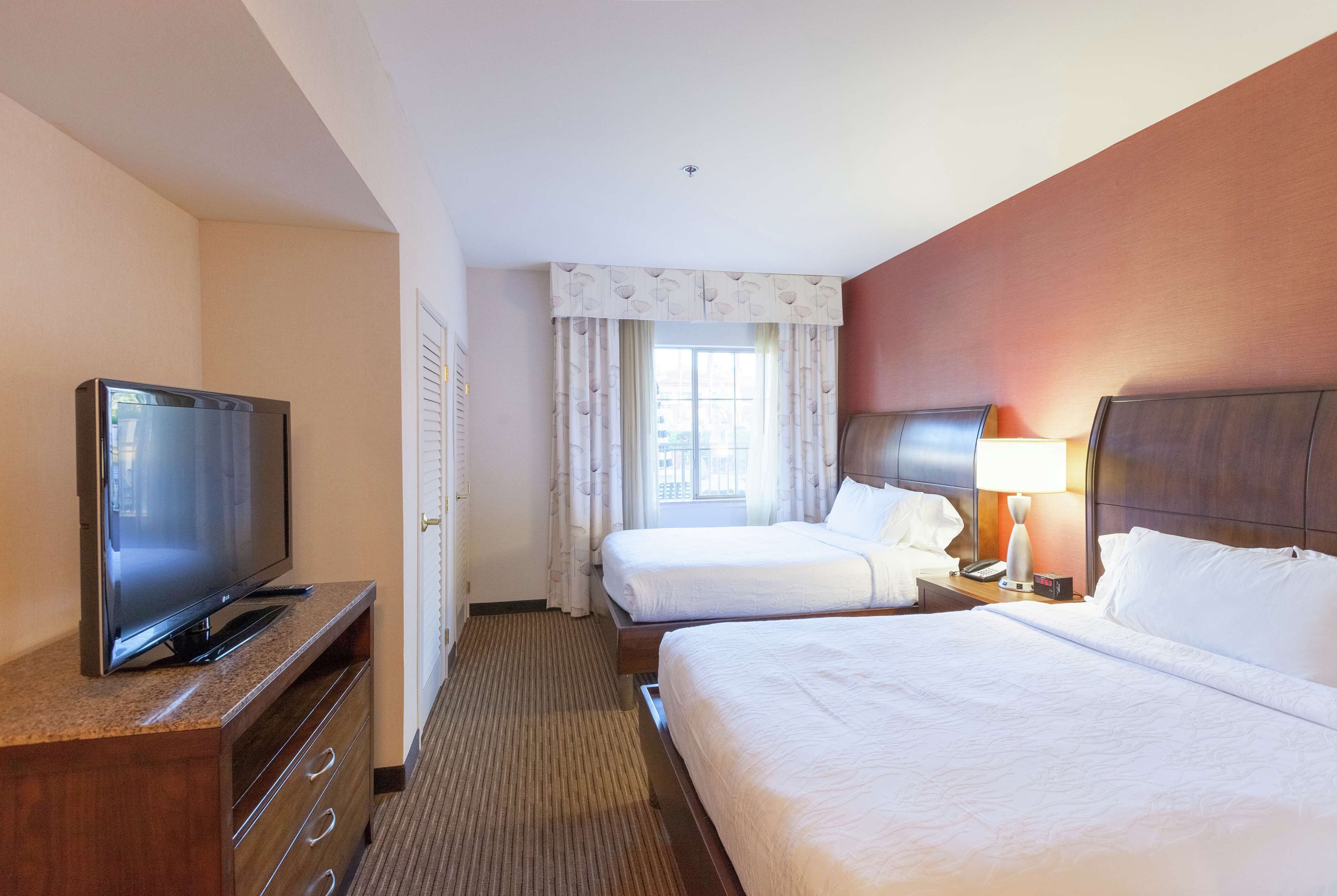 Hotelkamer met televisie, twee bedden, raam met gordijnen, lamp en houten hoofdeinde.