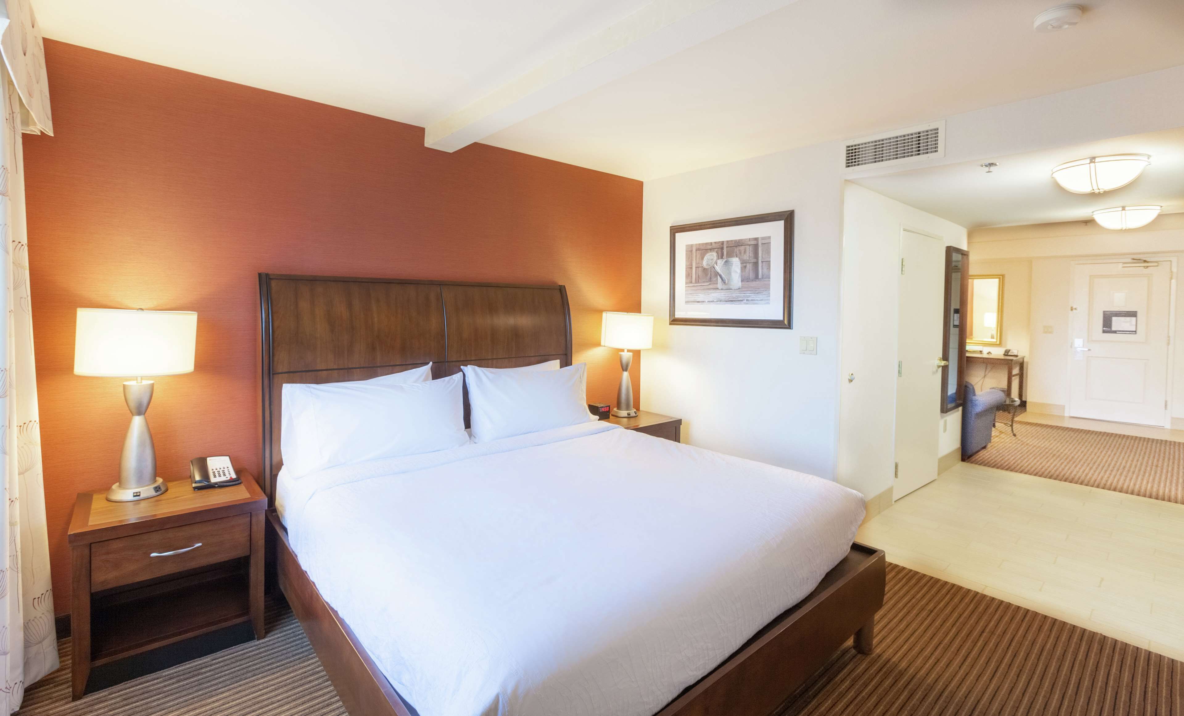 Una habitación de hotel con una cama grande, dos mesitas de noche con lámparas, un cuadro enmarcado en la pared y una puerta que da a otra habitación.