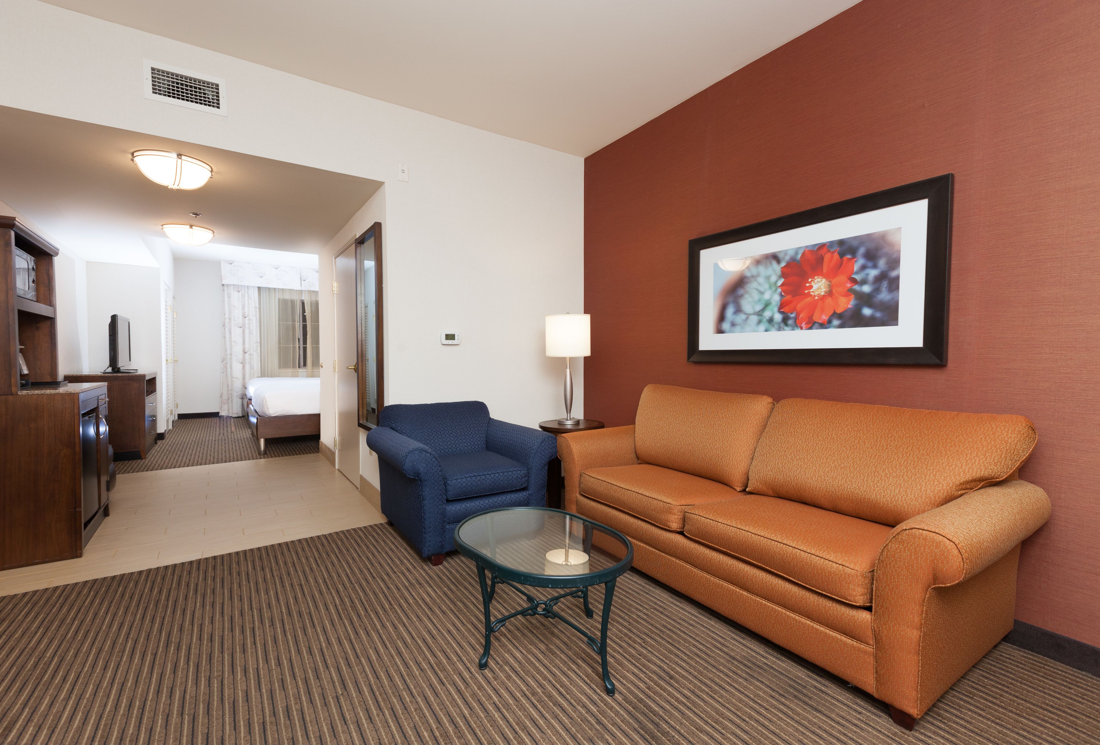 A hotel room with a sofa, a glass coffee table, a lamp, a framed picture on the wall, and a view into the adjacent room.