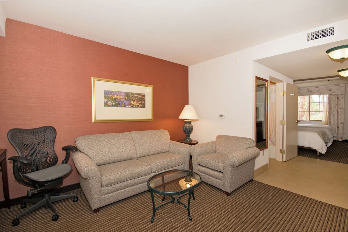 A hotel room with a two-seater sofa, a single-seater armchair, a glass coffee table, a lamp, and a bed with a window view.