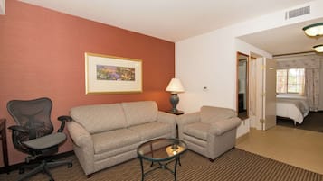 A hotel room with a two-seater sofa, a single-seater armchair, a glass coffee table, a lamp, and a bed with a window view.