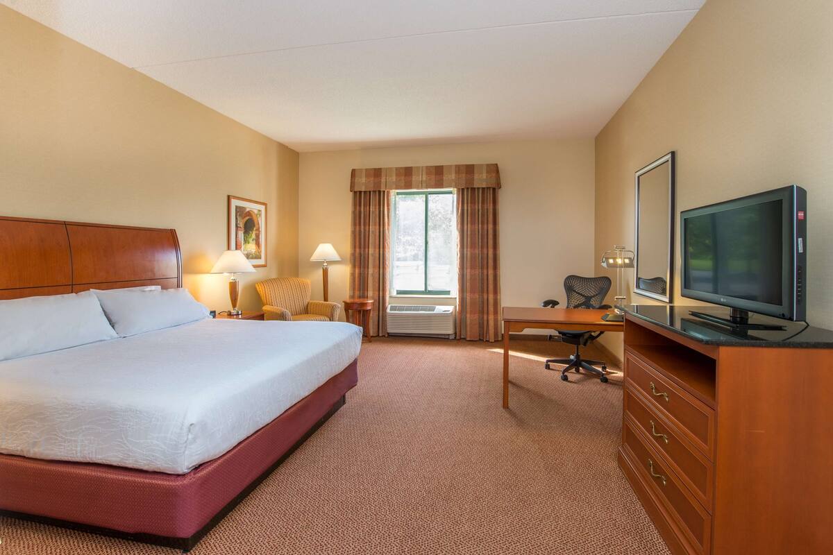 A hotel room with a large bed, a desk with a chair, a television, and a window with curtains.
