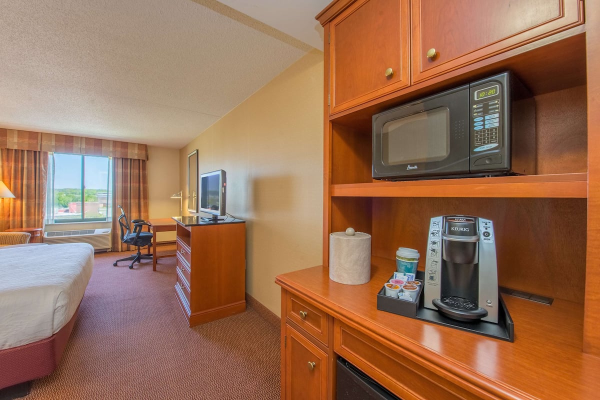 A hotel room with a bed, desk, chair, microwave, coffee maker, and a TV.