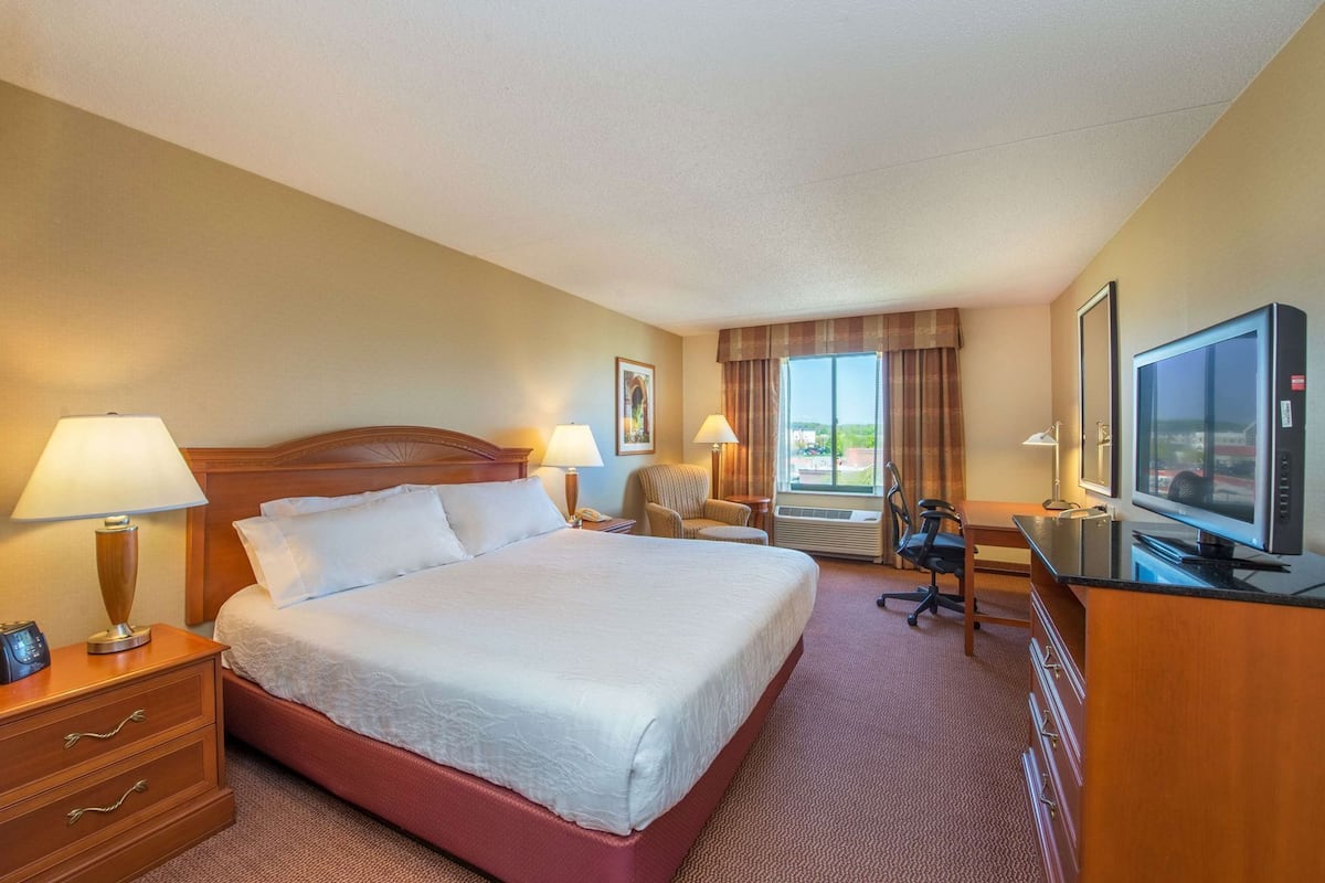 A hotel room with a large bed, two bedside tables with lamps, a desk with a computer, a TV, and a window with curtains.