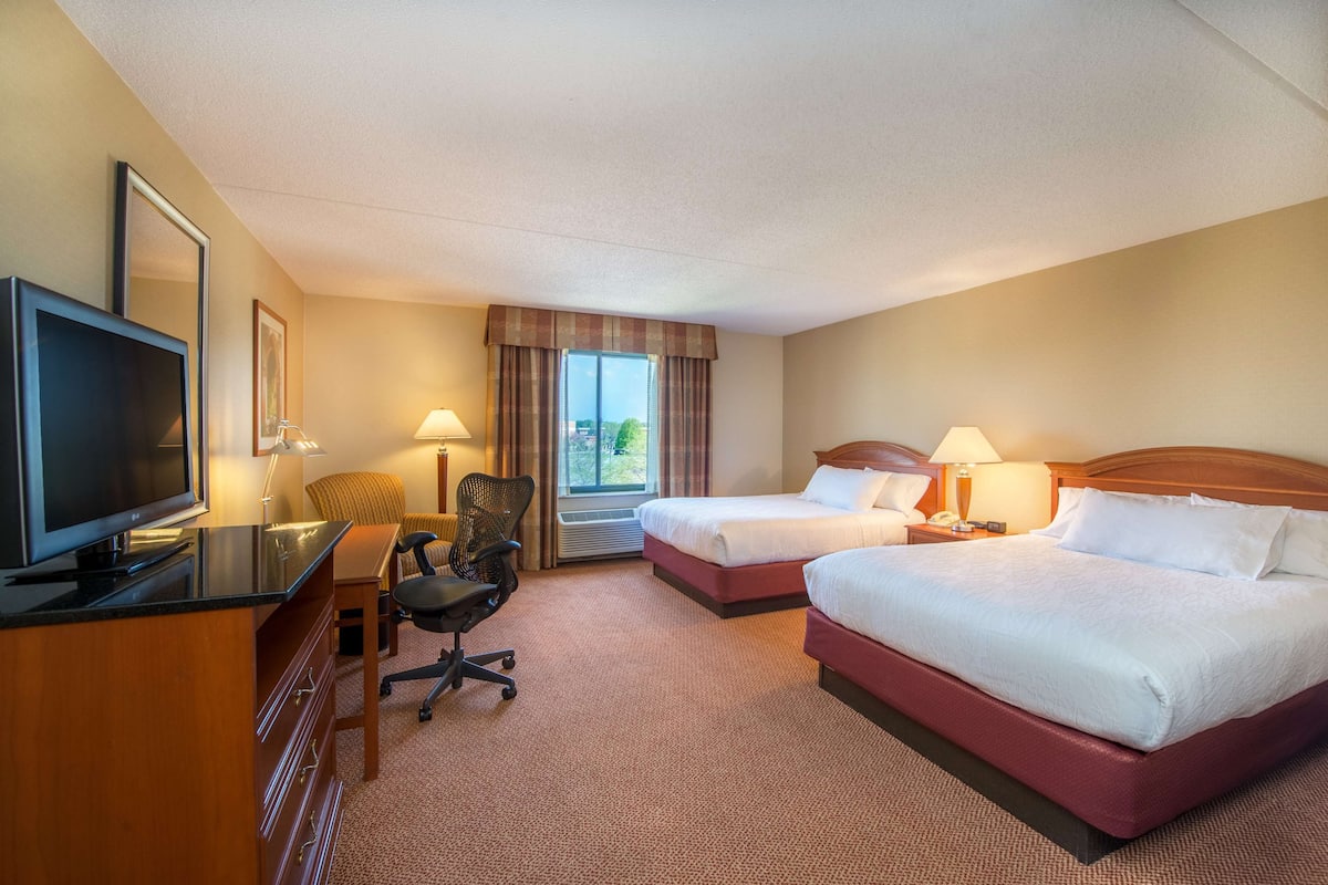 A hotel room with two beds, a desk with a TV, a chair, and a window with curtains.