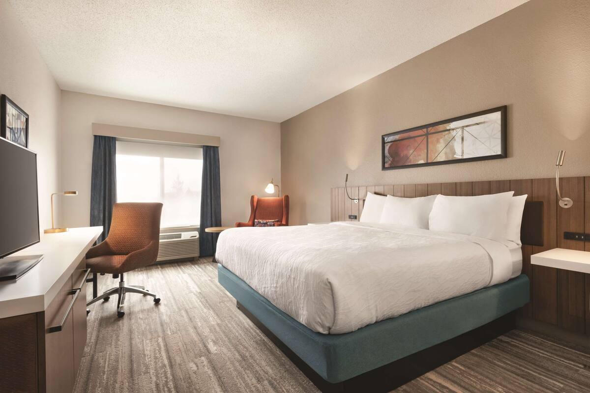 A hotel room with a large bed, a desk with a chair, a TV, and a window with curtains.