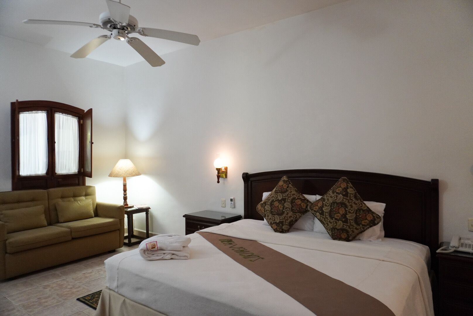 Una camera d'albergo con un letto, un divano, una ventola a soffitto, un comodino, una lampada e una finestra con tende.