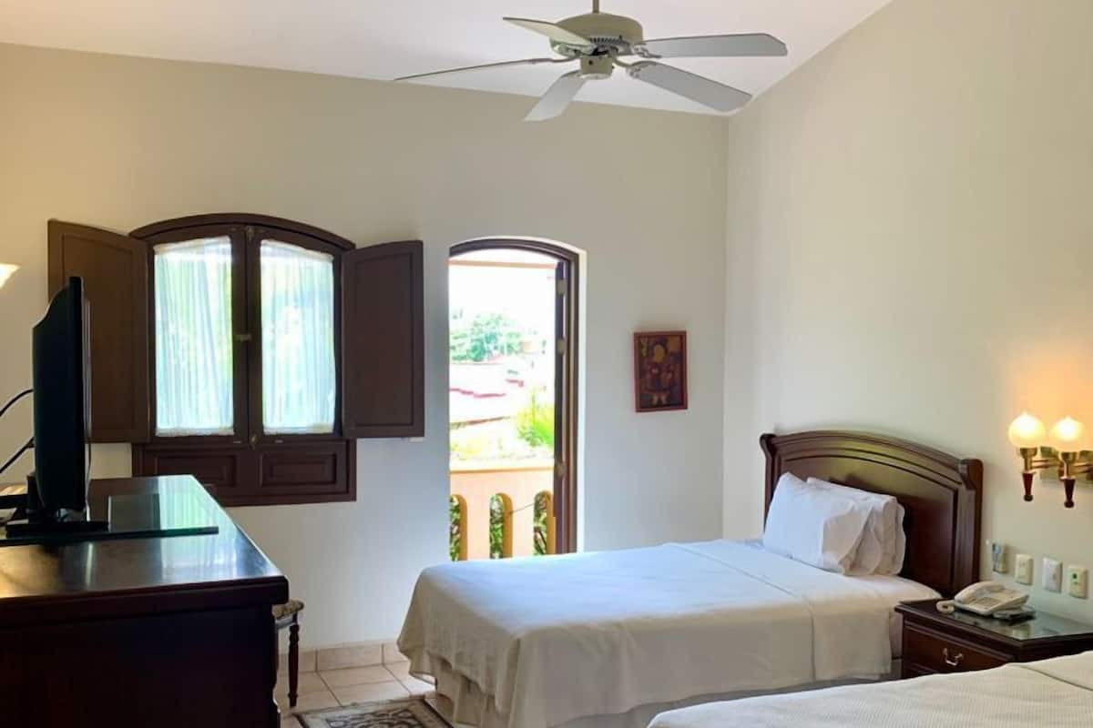 Quarto de hotel com duas camas, uma escrivaninha, ventoinha no teto e vista para o exterior.