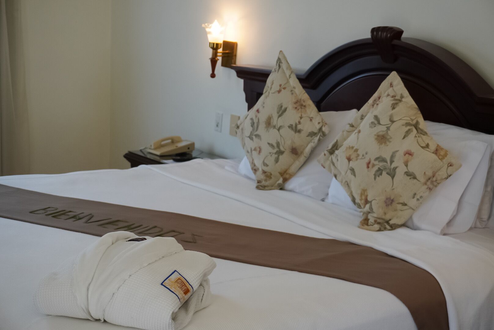 A neatly made bed with floral pillows, a brown bedspread, and a folded robe on the side.