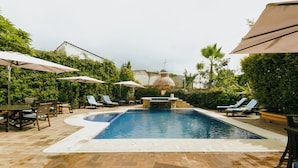 Piscina con sillas de descanso y sombrillas, rodeada de vegetación y un patio de ladrillo.