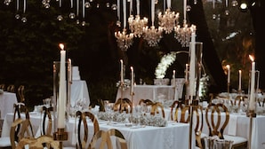 Un área de comedor al aire libre con manteles blancos, detalles dorados en las sillas y mesas iluminadas con velas.