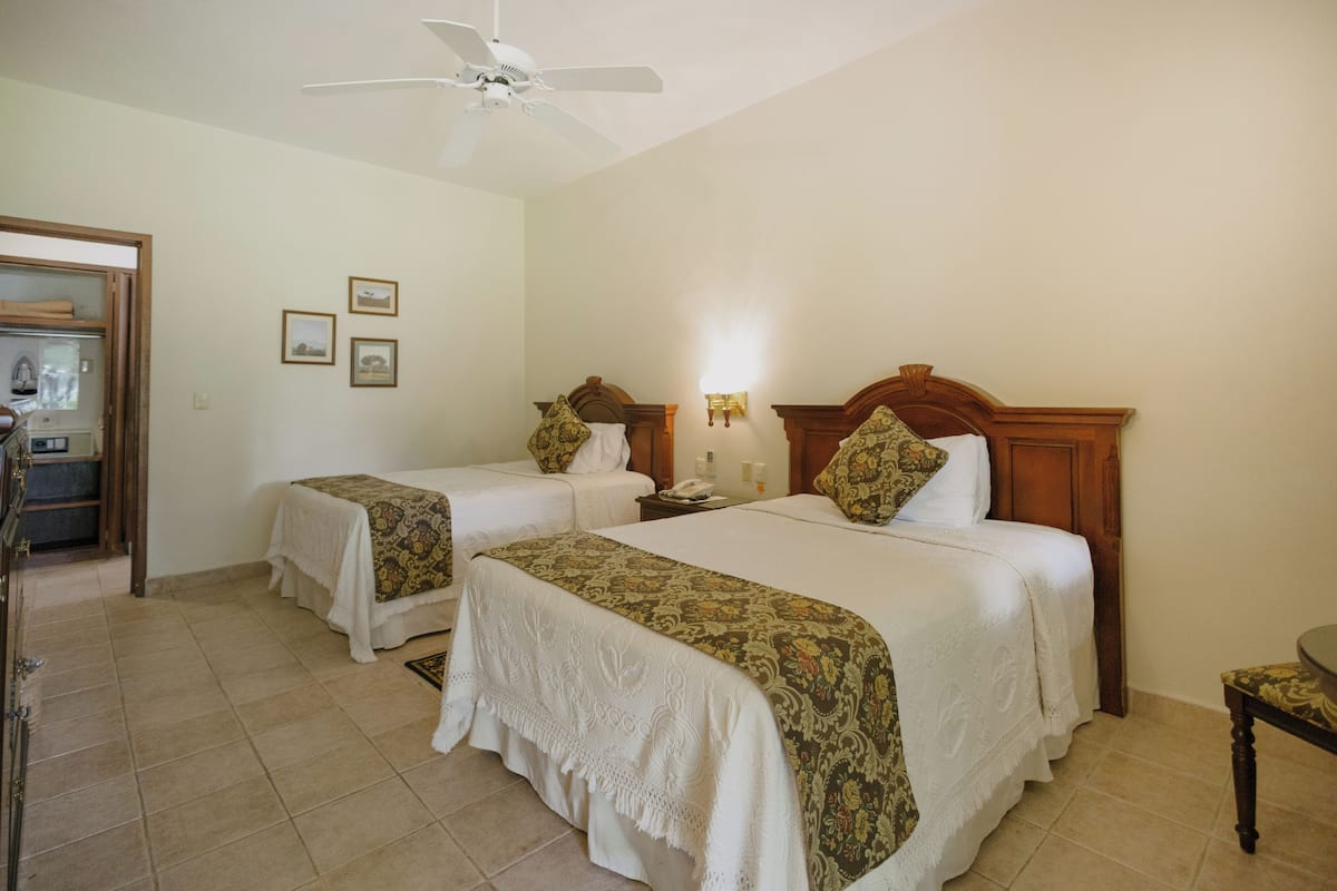Quarto de hotel com duas camas, um ventilador de teto, uma mesinha e uma cadeira.
