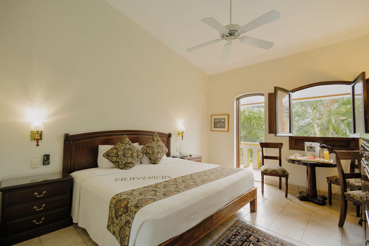 Quarto de hotel com uma cama, escrivaninha, cadeiras, ventoinha no teto e vista para o verde pela janela.