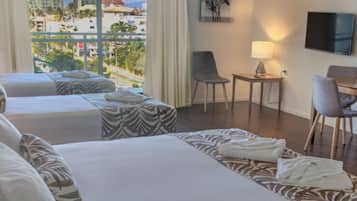 A hotel room with two beds, a desk, chairs, a TV, and a view of the city.