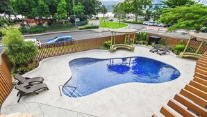 Piscina con sillas de descanso y un pérgola de madera, rodeada por una valla y follaje.