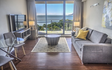 A modern living room with a sofa, a glass coffee table, and a TV. It features a large window with a view of the sea.