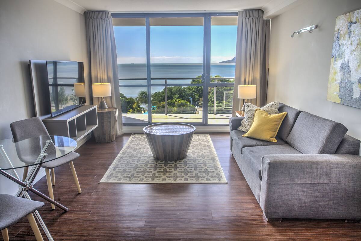 Een moderne woonkamer met een bank, een glazen salontafel en een televisie. Er is een groot raam met uitzicht op zee.