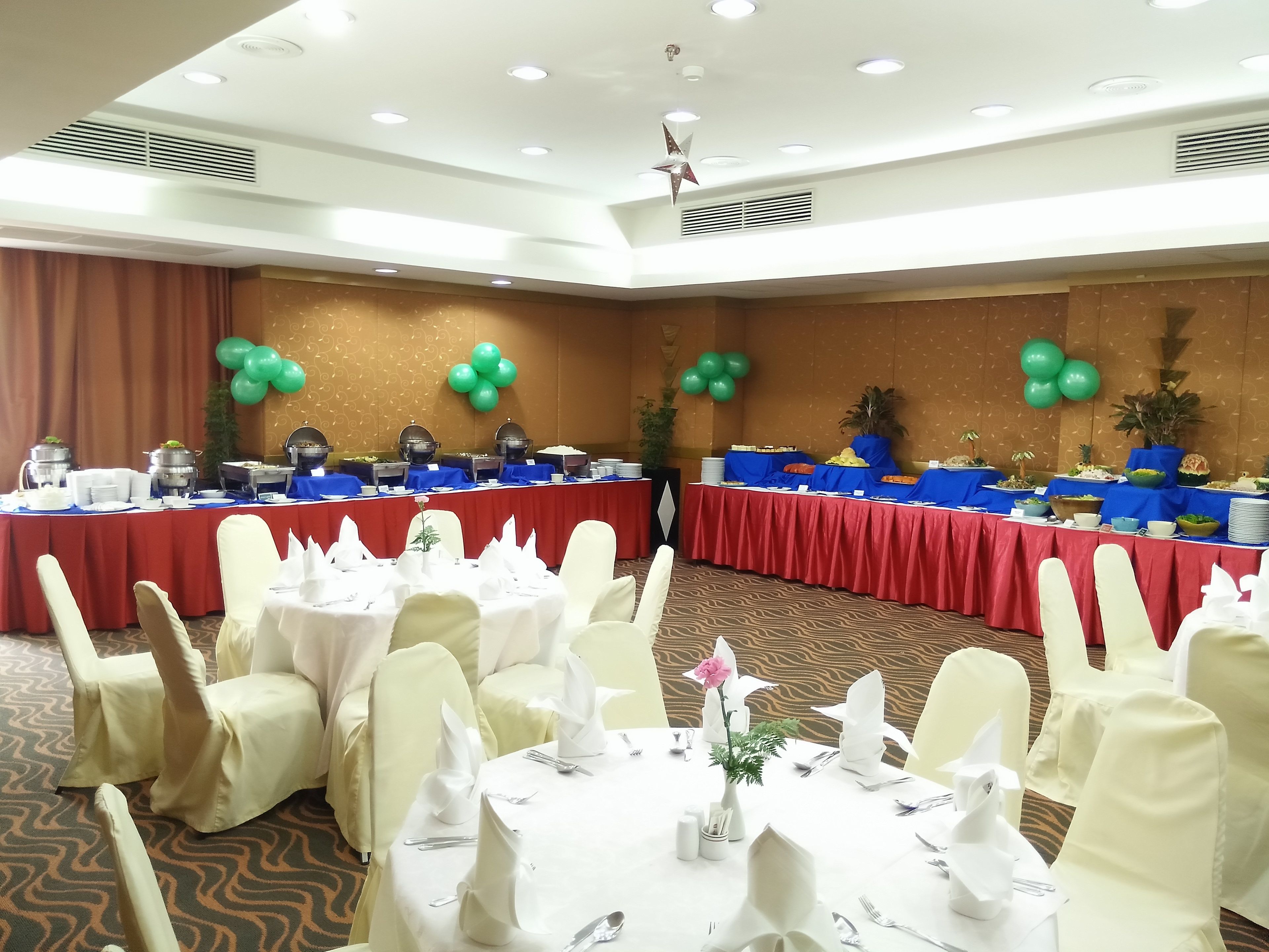 A banquet hall with round tables set for dining, a buffet line with various food stations, and green balloons on the walls.
