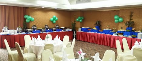 A banquet hall with round tables set for dining, a buffet line with various food stations, and green balloons on the walls.