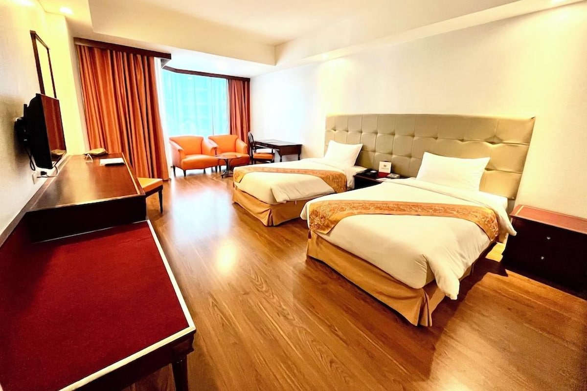 A hotel room with two beds, a desk, a chair, a TV, and a large window with curtains.