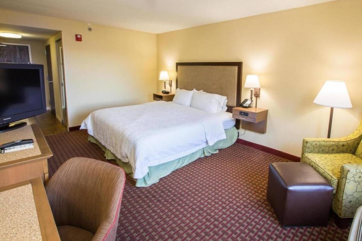 A hotel room with a bed, two bedside tables with lamps, a television, a chair, and a small ottoman.