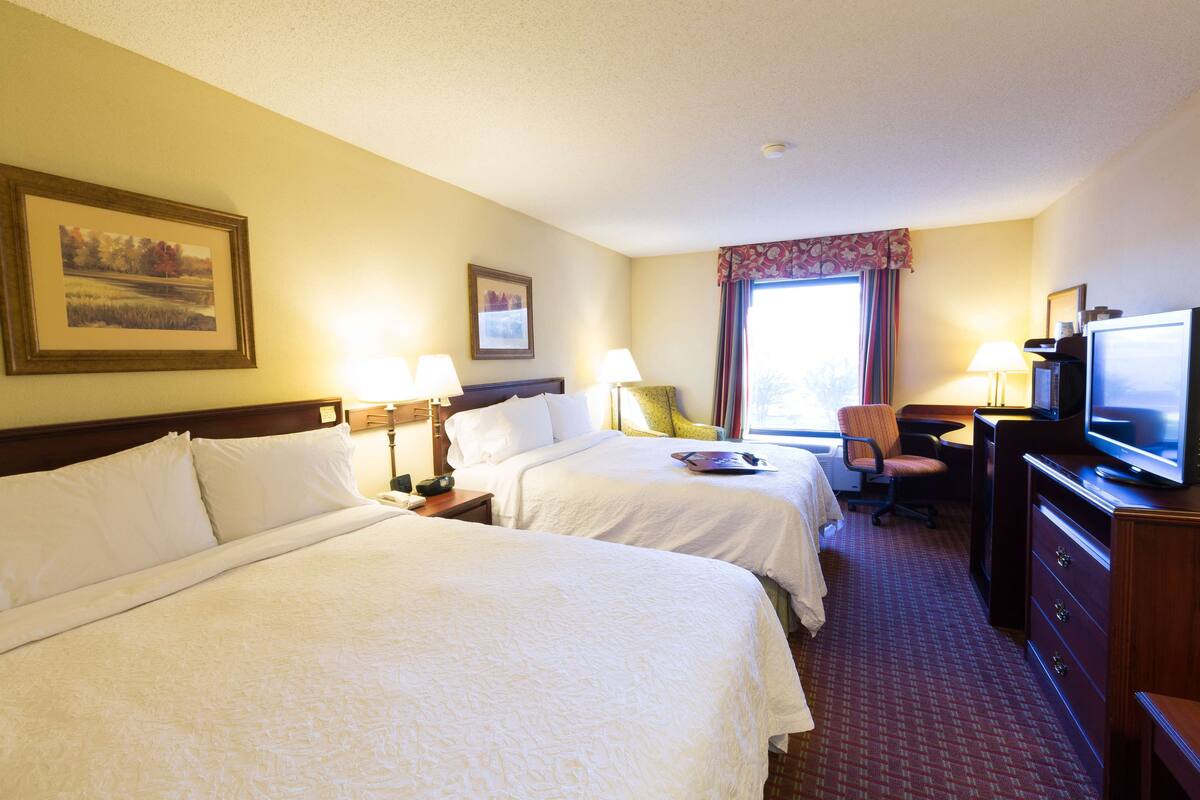A hotel room with two beds, a desk with a television, a chair, and a desk lamp.