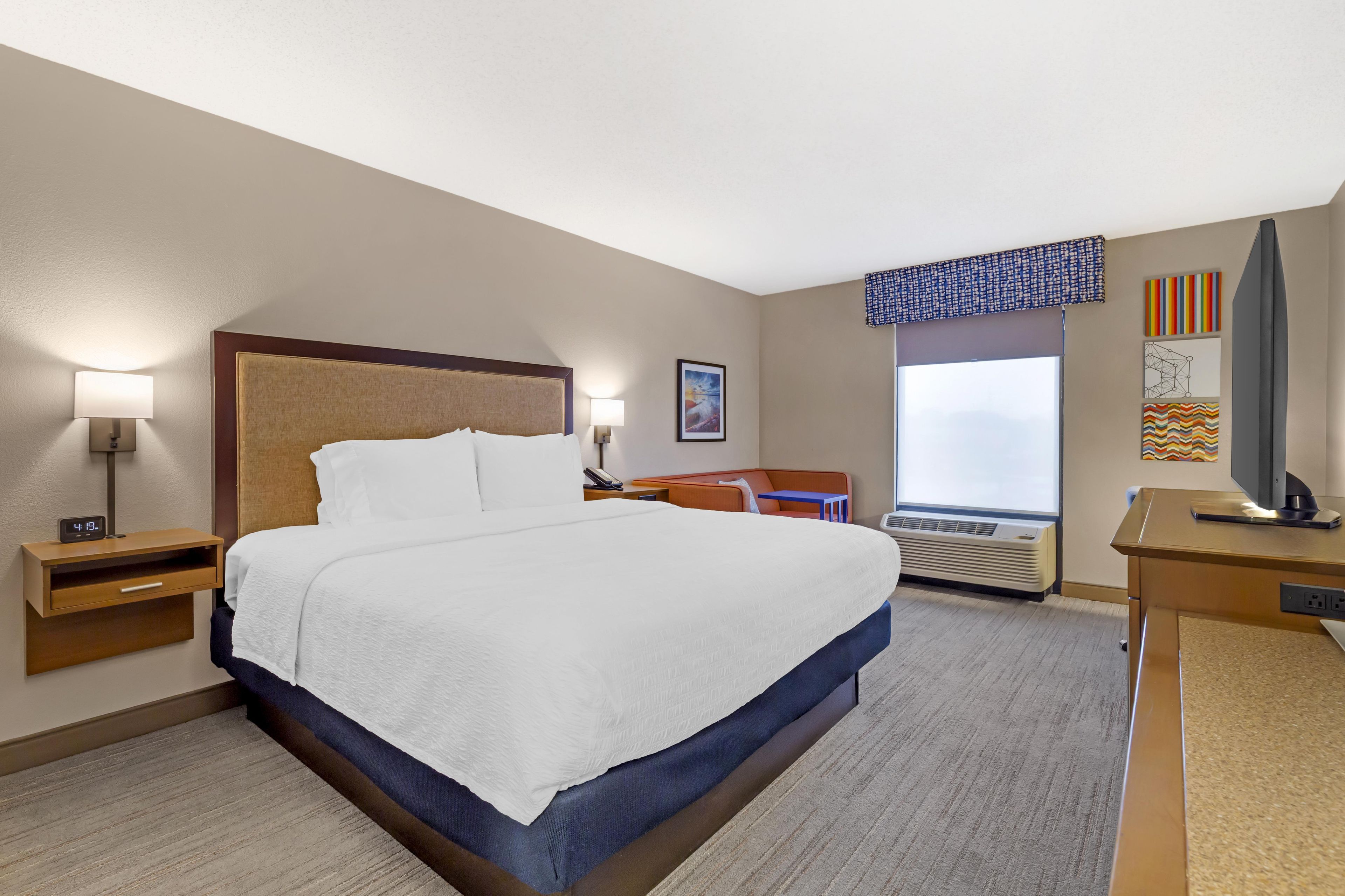 A hotel room with a large bed, bedside lamps, a desk, a chair, a window with a view, and artwork on the walls.