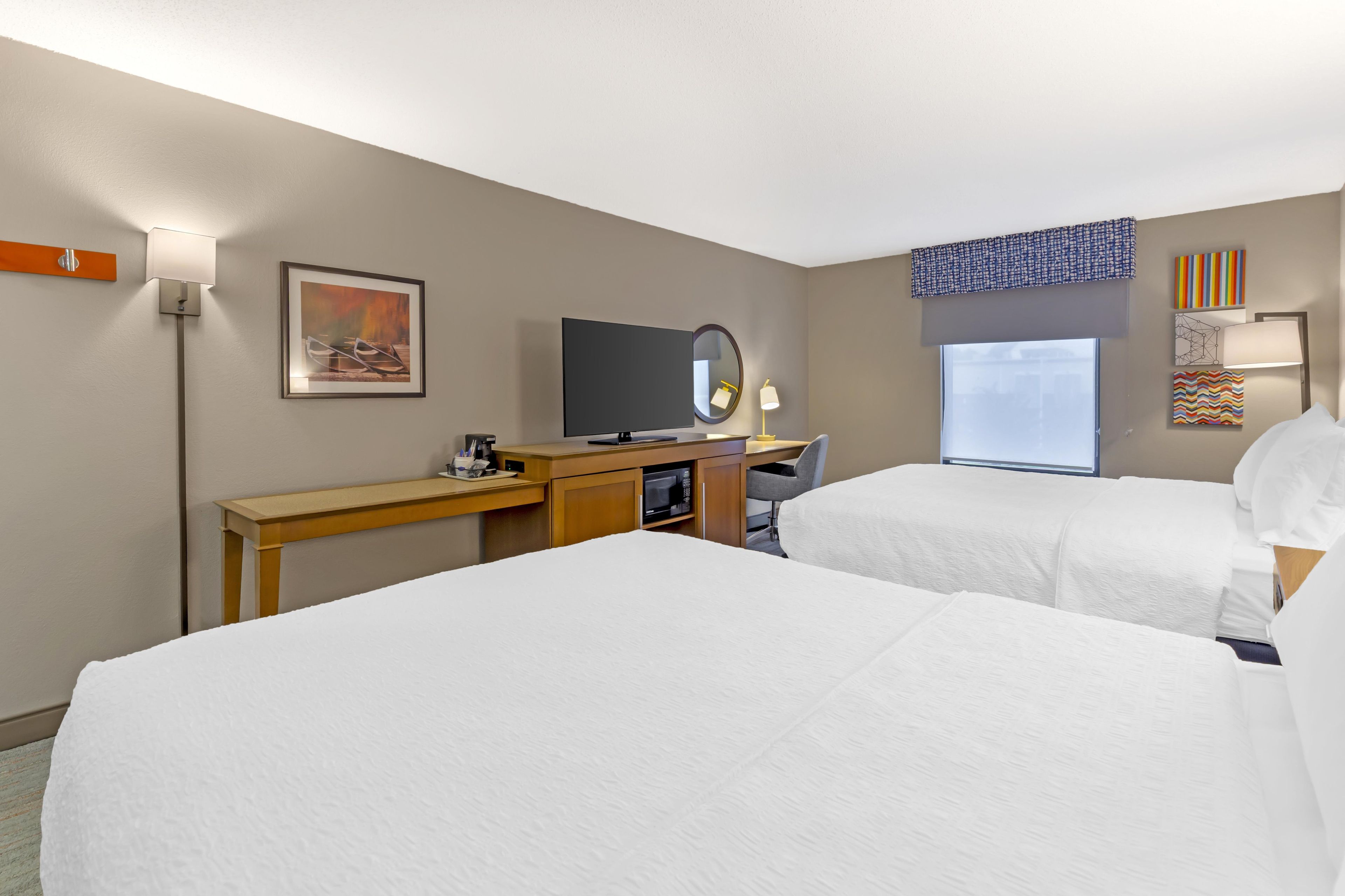 A hotel room with two beds, a desk with a TV, a mirror, and a window with blinds.