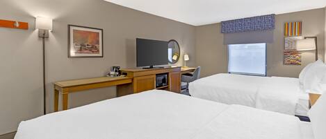 A hotel room with two beds, a desk with a TV, a mirror, and a window with blinds.