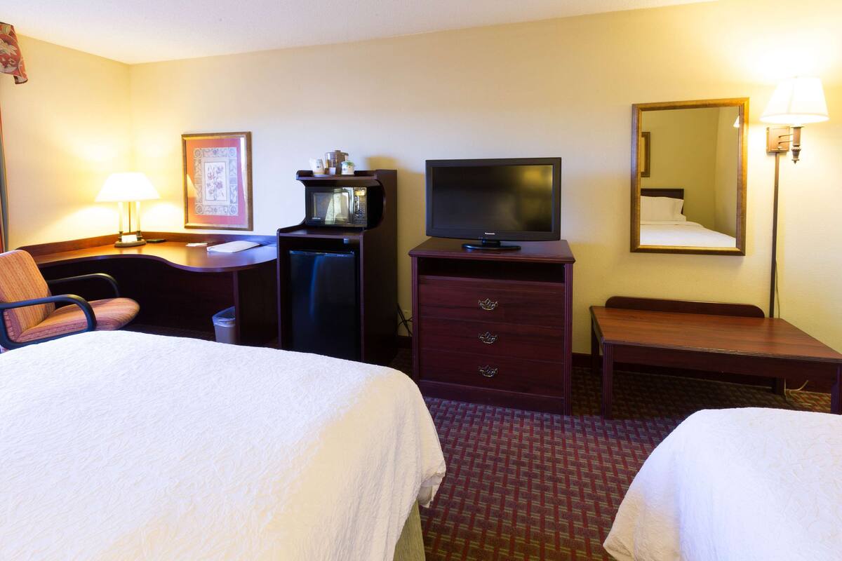 A hotel room with two beds, a desk with a lamp, a television, a microwave, and a dresser.