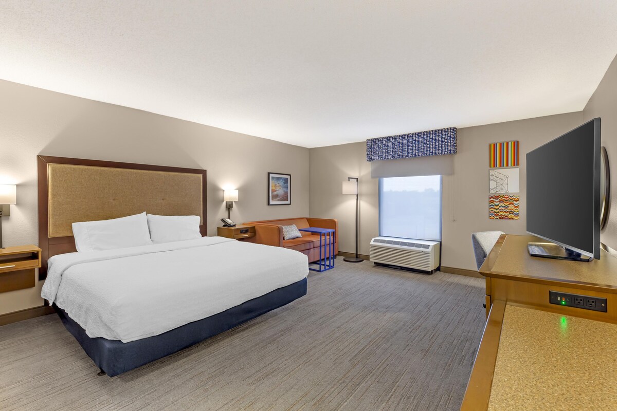 Habitación de hotel con una cama grande, un escritorio con televisor de pantalla plana, una lámpara de escritorio, una mesita de noche, una silla, un ventanal con vistas y un aire acondicionado de pared.