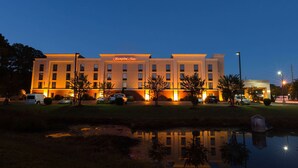 Un hotel de varios pisos con una fachada bien iluminada, un área de estacionamiento y un pequeño estanque al frente.