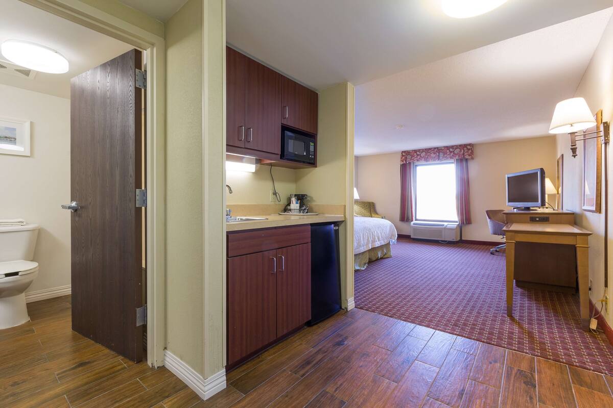 A hotel room with a wooden floor, a kitchenette with a microwave, a bed, a desk, and a TV.