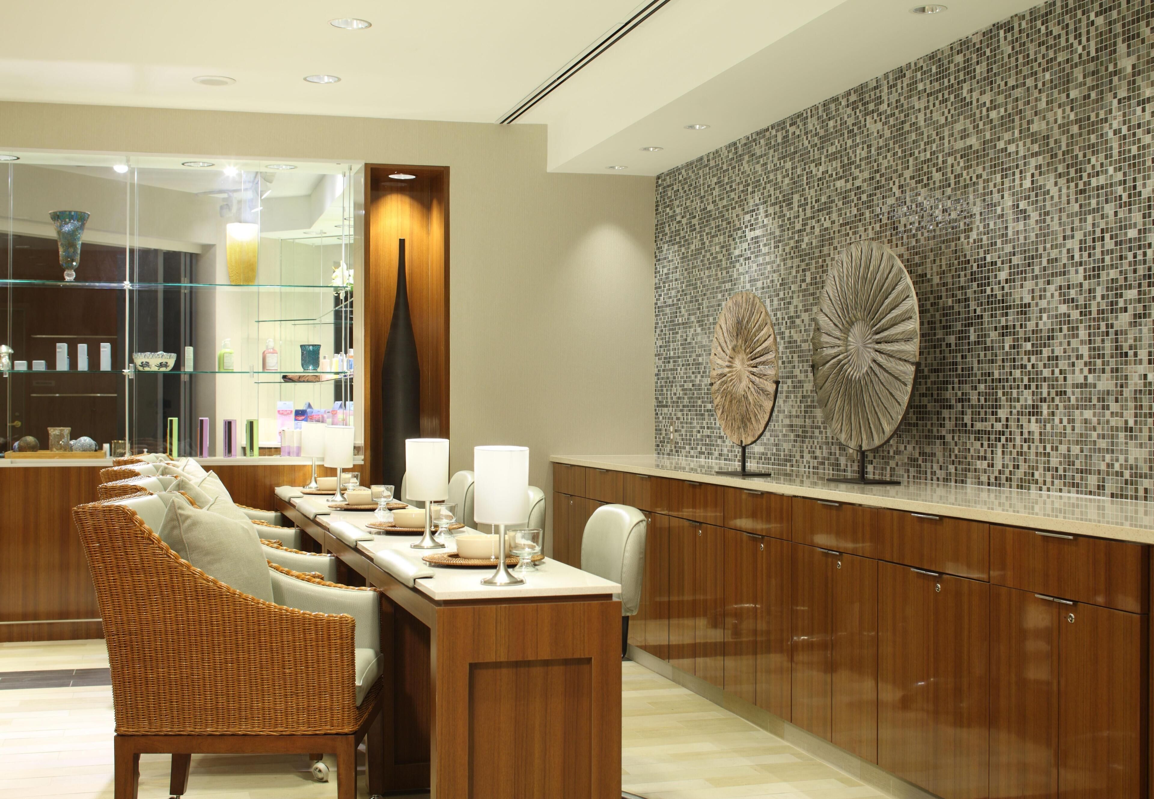A modern hotel lobby with a long dining table, wicker chairs, a glass display case, and a decorative wall with mosaic tiles.