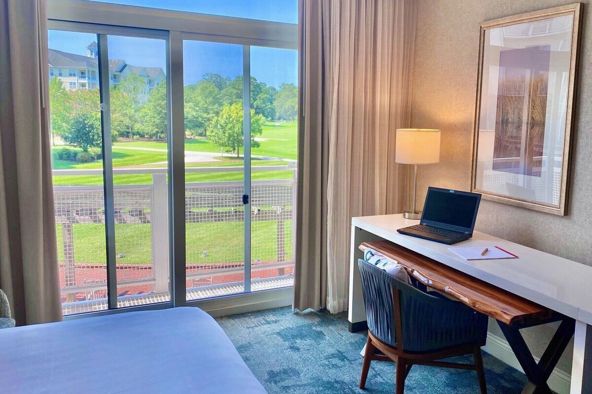 A hotel room with a bed, a desk with a laptop, a chair, a lamp, and a large window with a view of a golf course.