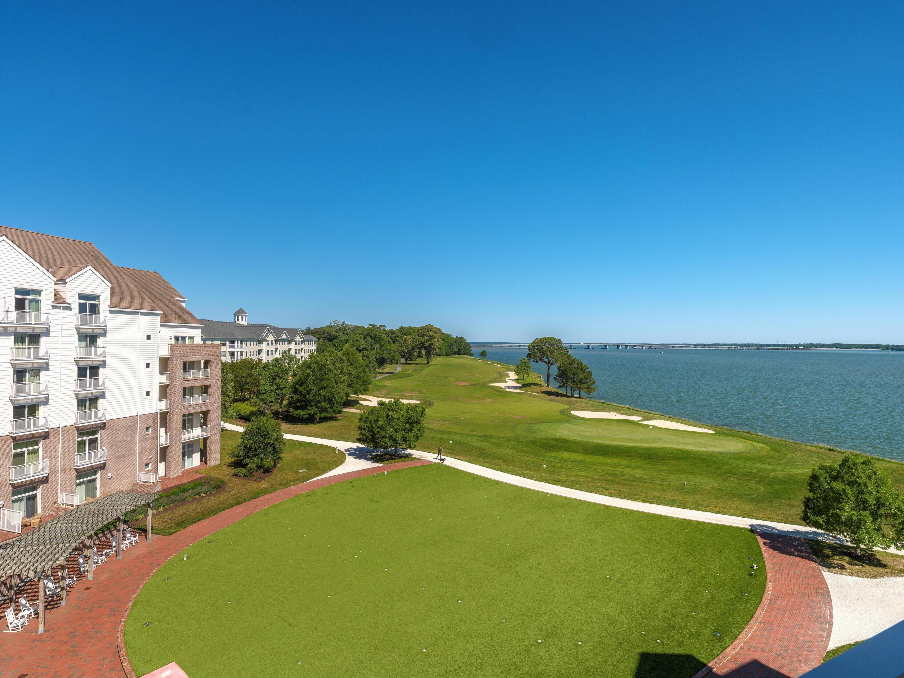 A golf course with a clubhouse, a large body of water, and a bridge in the background.
