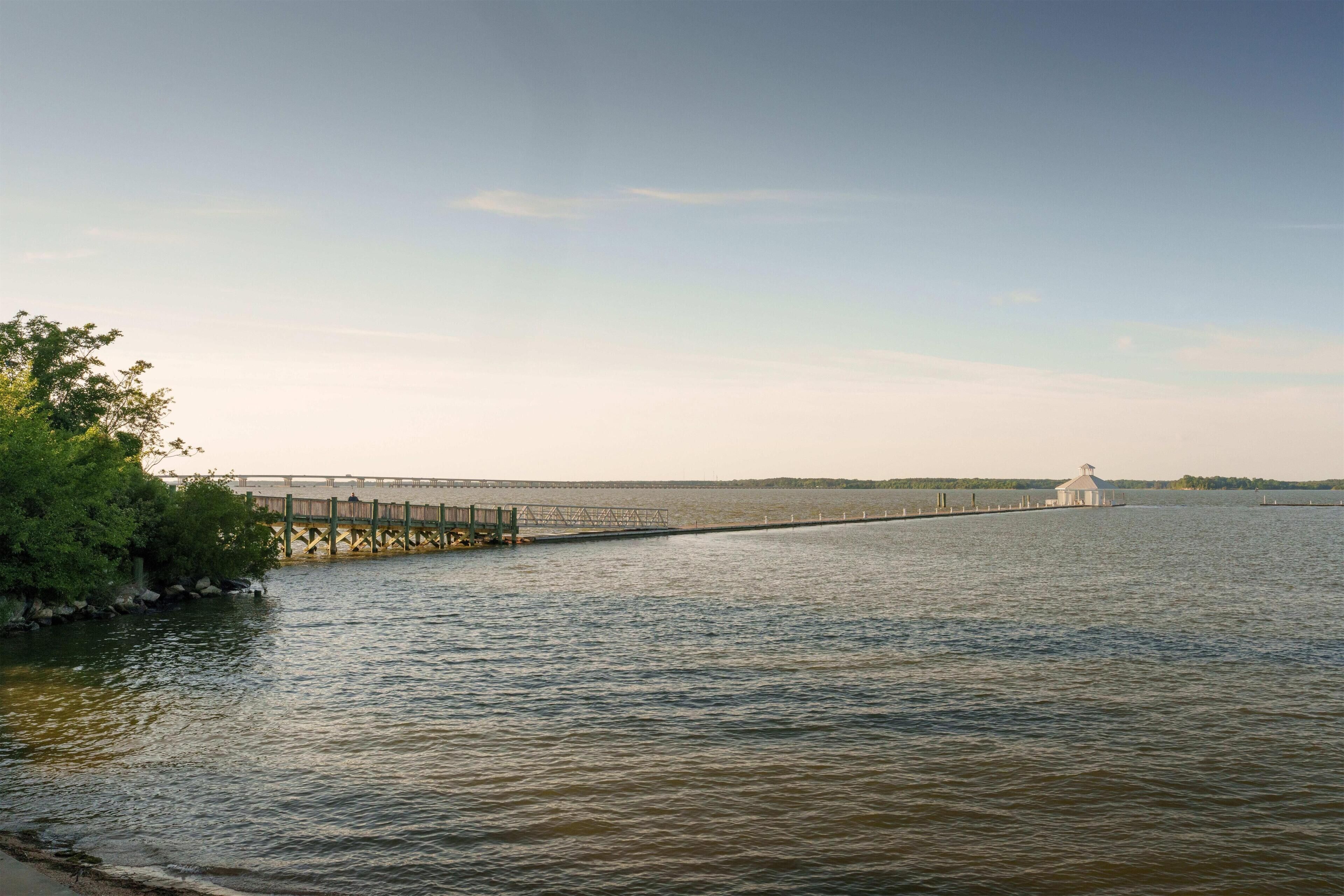 A calm river with a wooden pier extending into the water, a small white building at the end, and a forested area on the left.