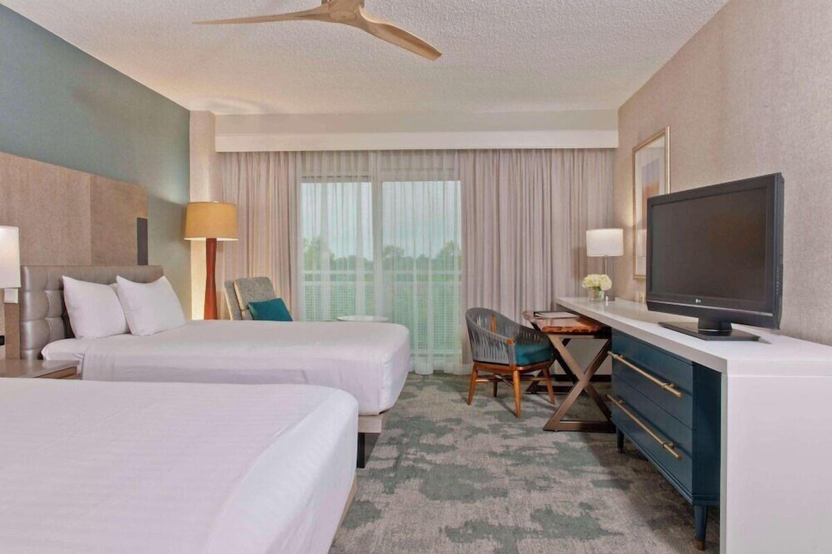 A hotel room with two beds, a desk with a TV, a chair, and a ceiling fan.