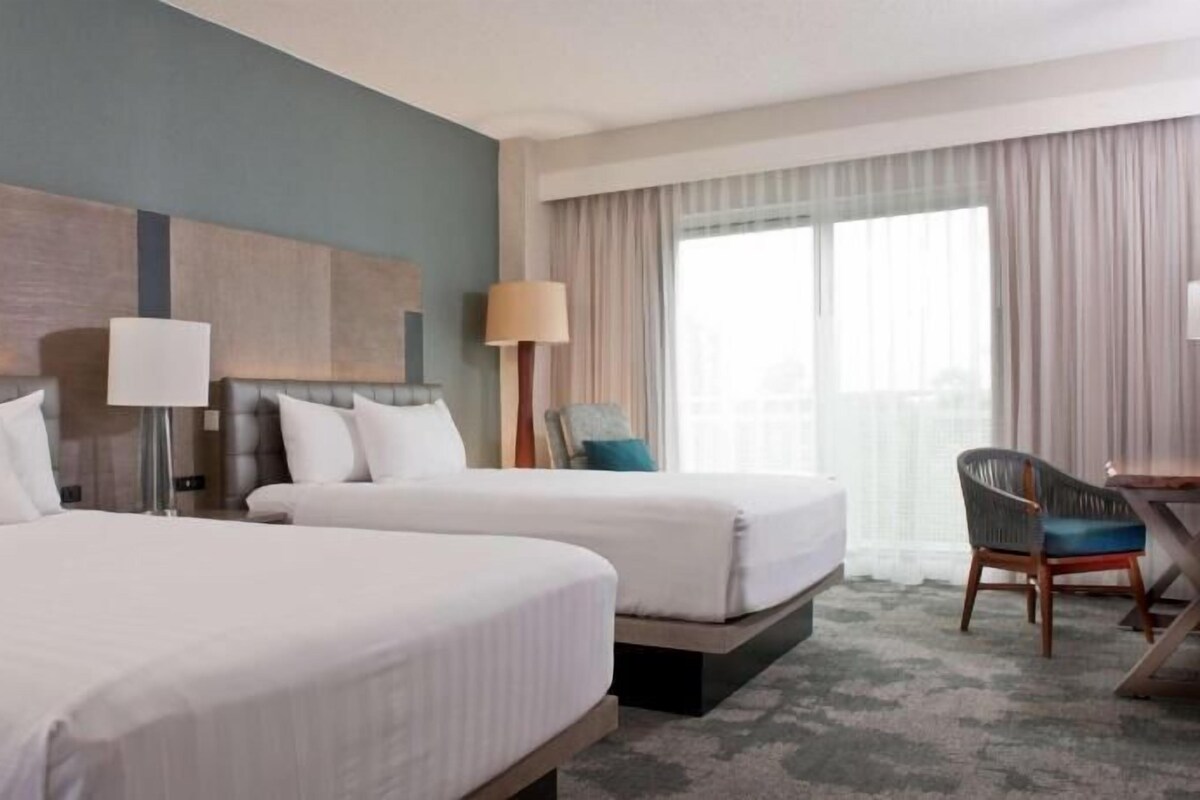 A hotel room with two beds, a desk, a chair, a lamp, and a large window with curtains.