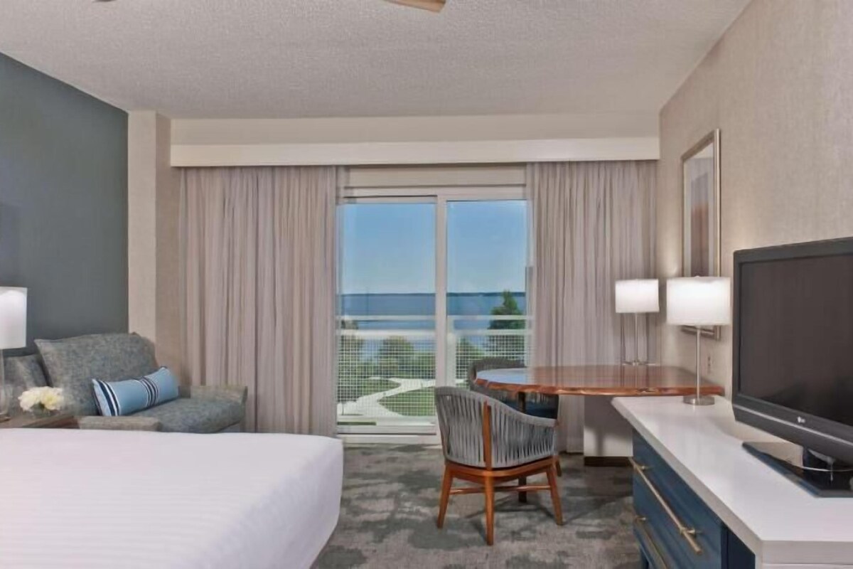 A hotel room with a bed, a desk with a TV, a chair, a lamp, and a view of the outdoors.