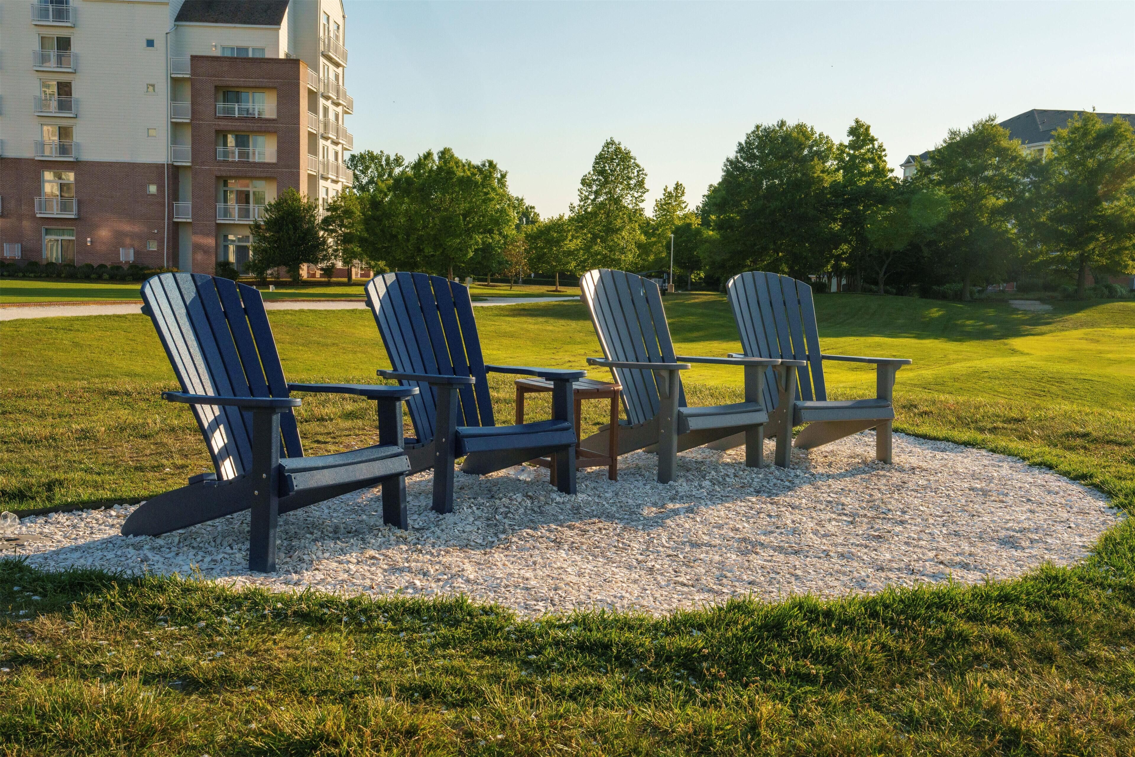 Three Adirondack chairs arranged on a gravel area with grass, surrounded by a building with multiple windows and balconies.