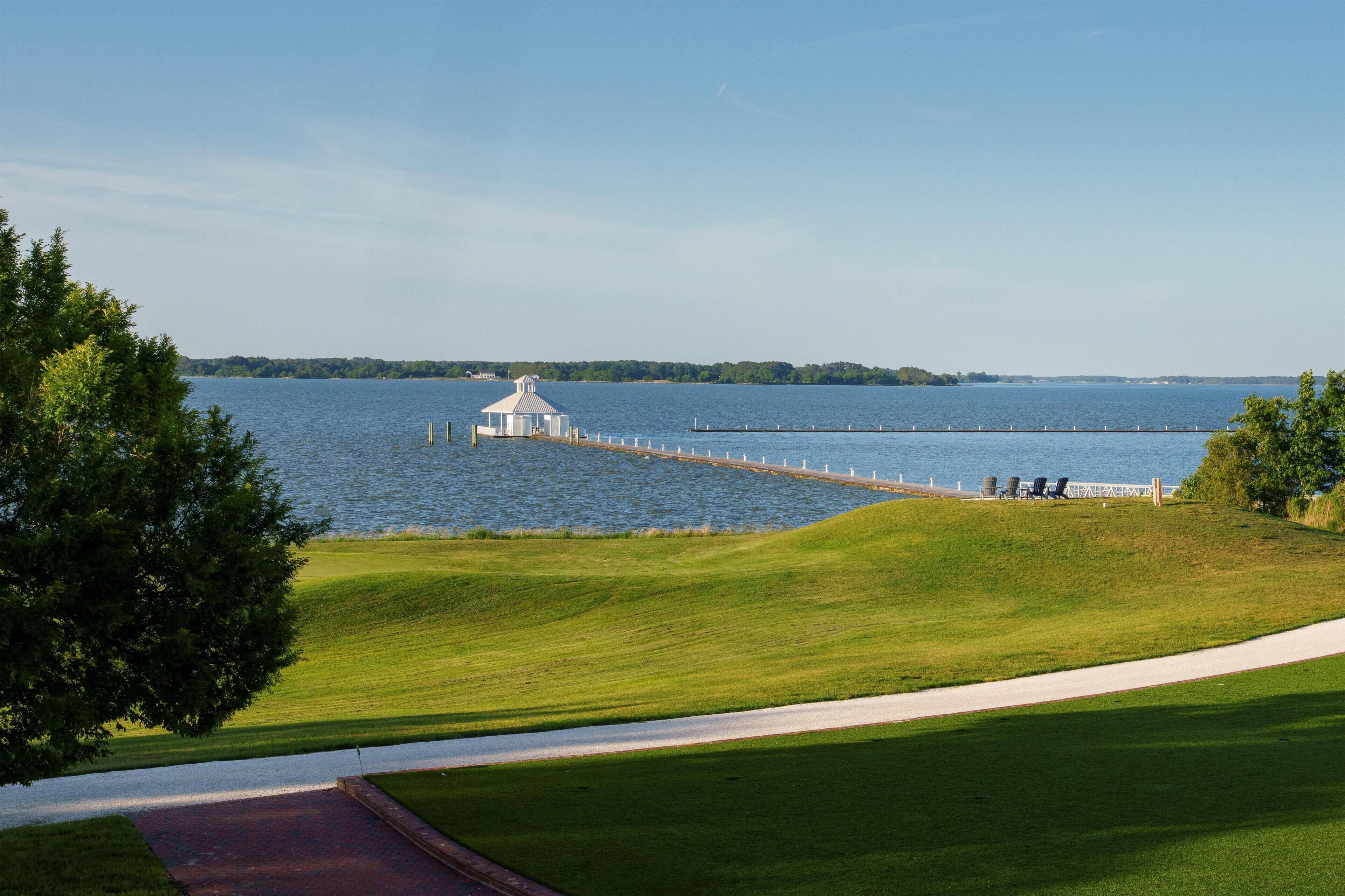 A grassy area with a dock extending into a body of water, a gazebo, and a distant shoreline.