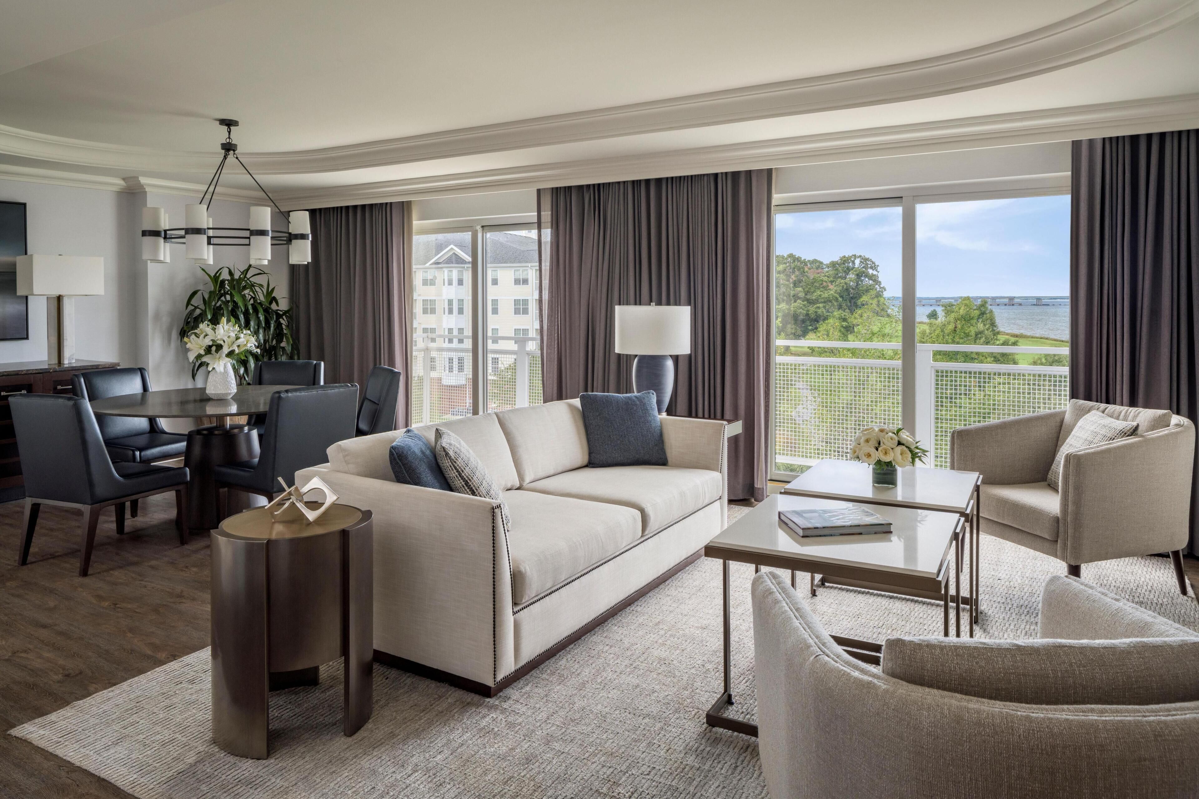 Habitación de hotel moderna con sofá, sillones, zona de comedor y vistas a un puente y vegetación.