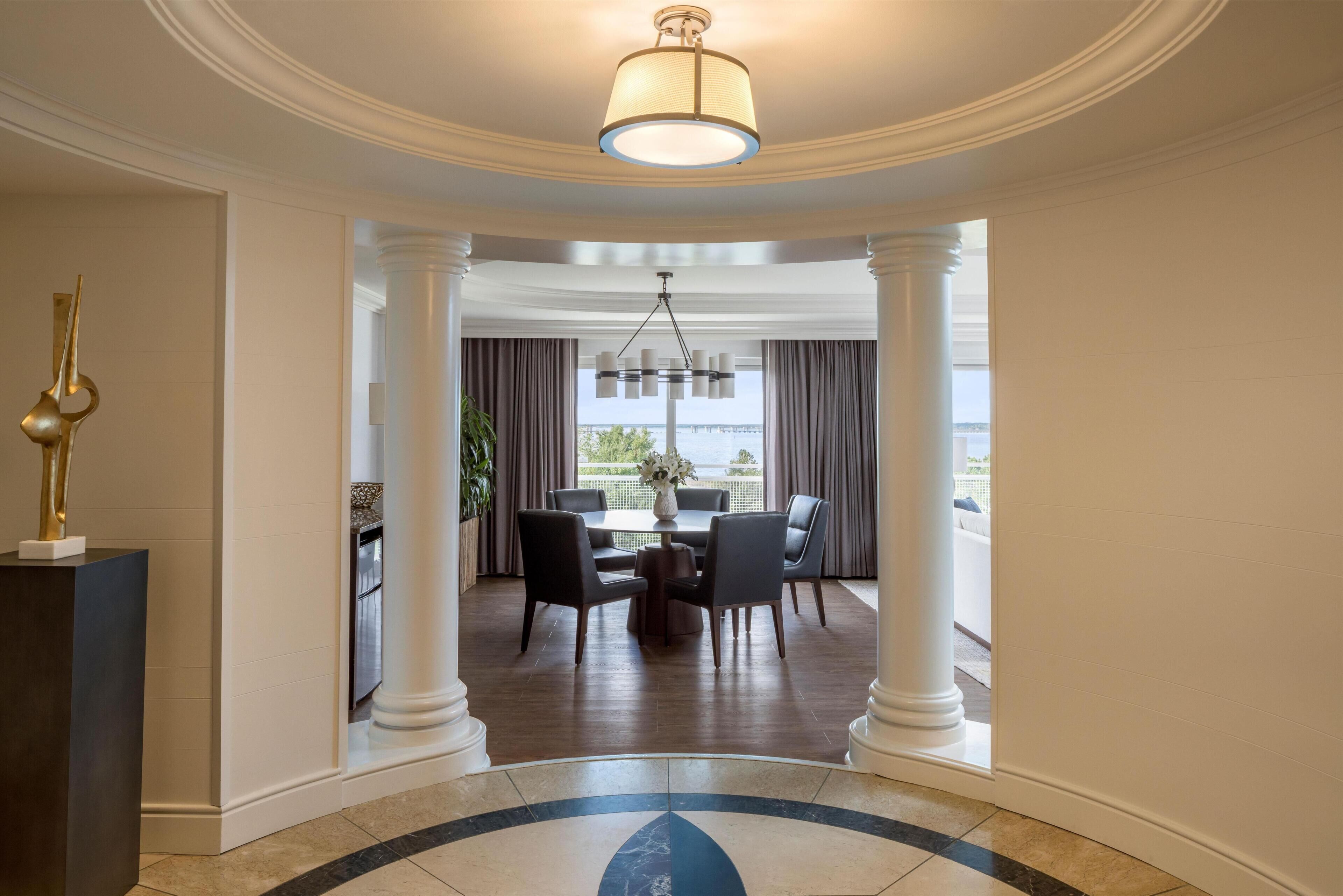 A hotel lobby with a dining area, white columns, a chandelier, and a view of the sea.