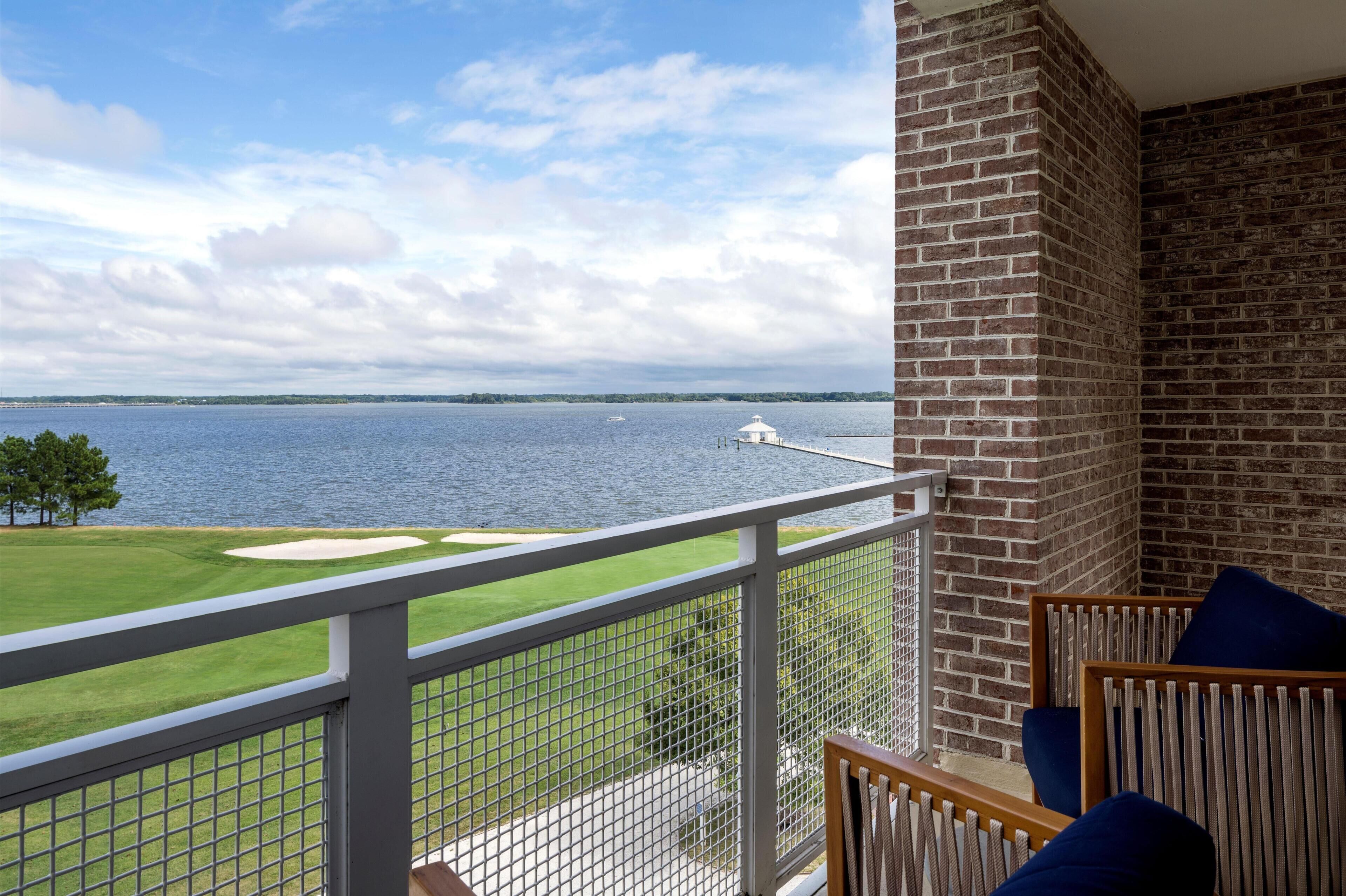 A balcony with a view of a lake, a white building on the water, and a grassy area with a fence.